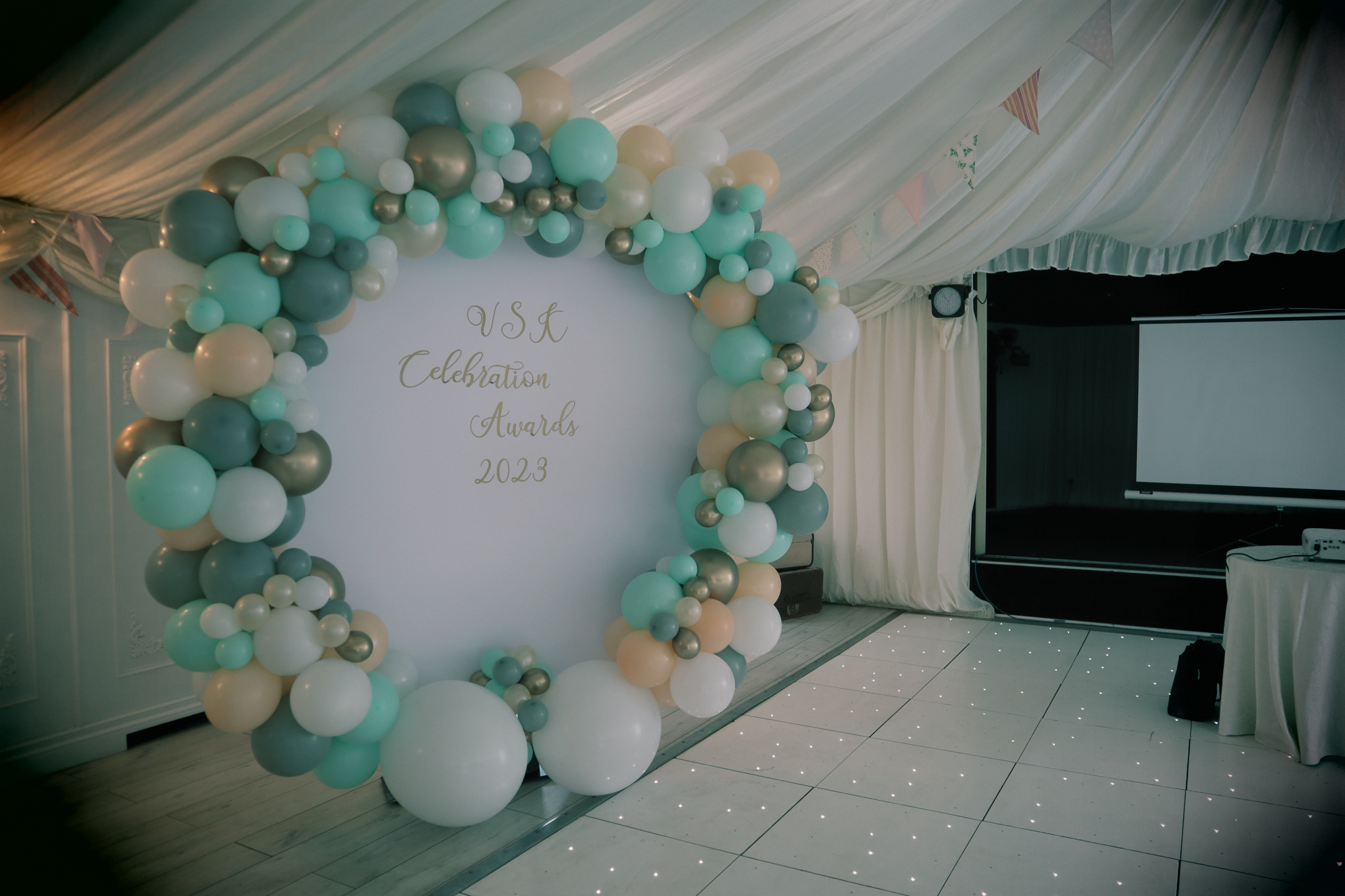 Awards room with a projector, tables and a backdrop with 'VSK Celebration Awards 2023'  framed in green, white and gold balloons